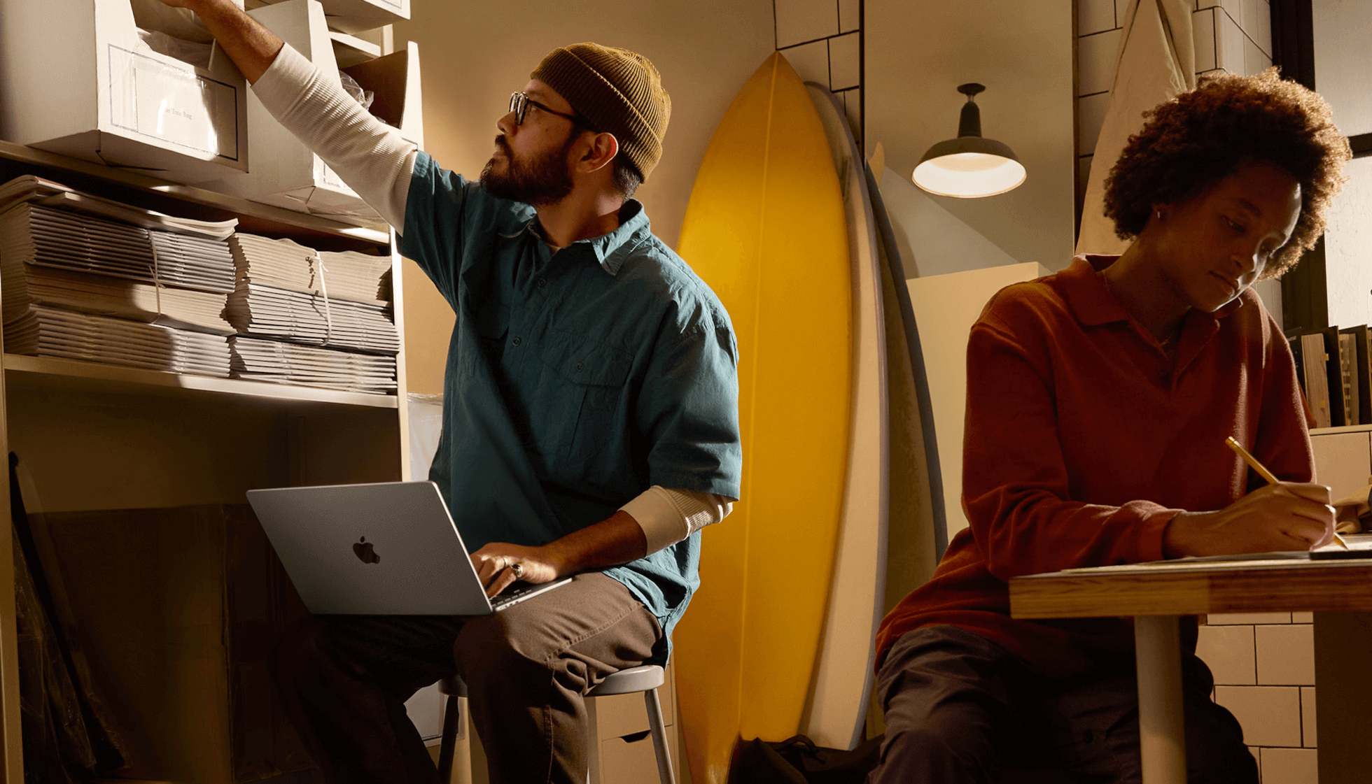 Person in work environment seated on stool using MacBook Air unplugged on their lap while reaching for box on high shelf with other hand
